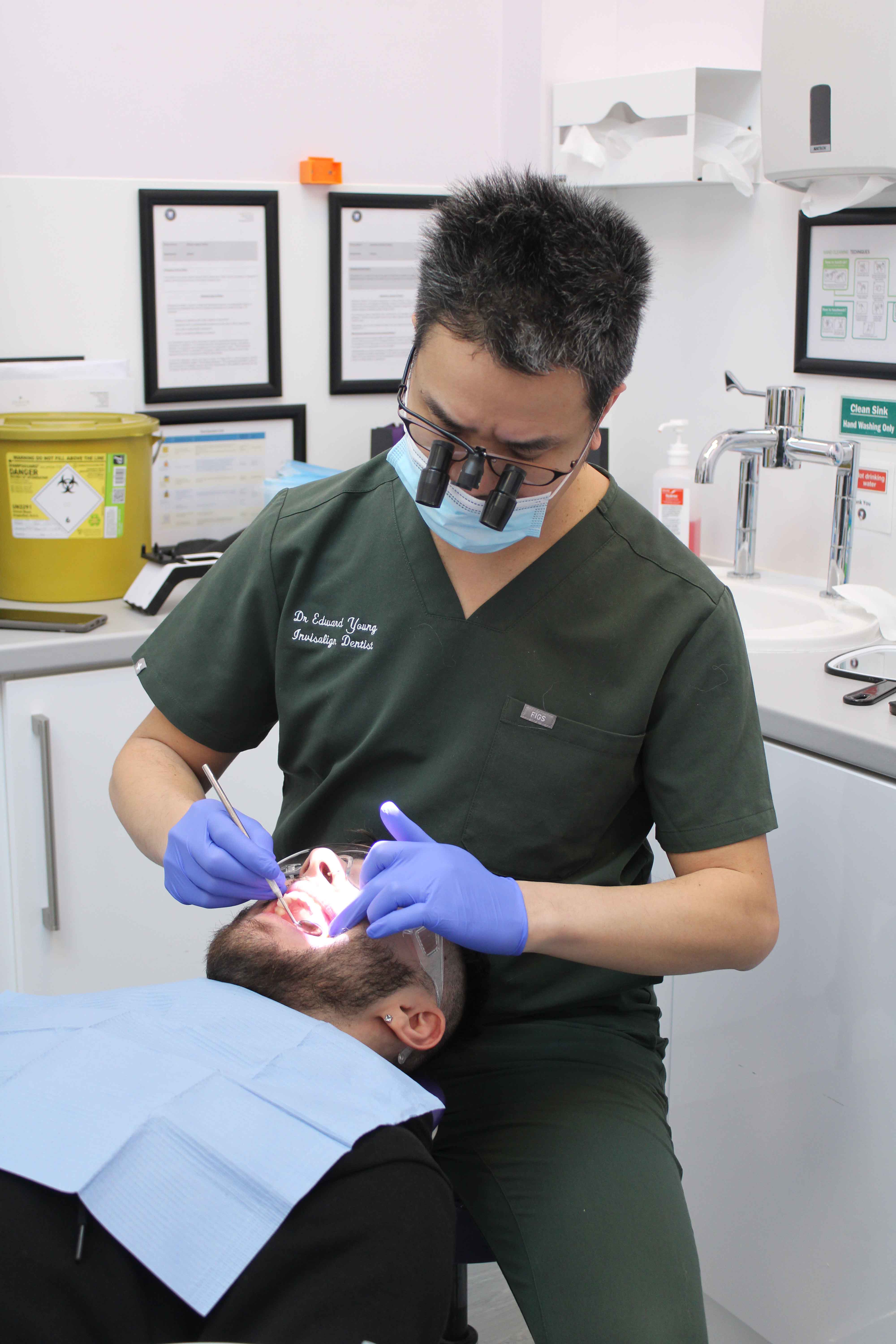 A dental professional wearing blue gloves examines a reclining patient’s mouth under a bright dental light.